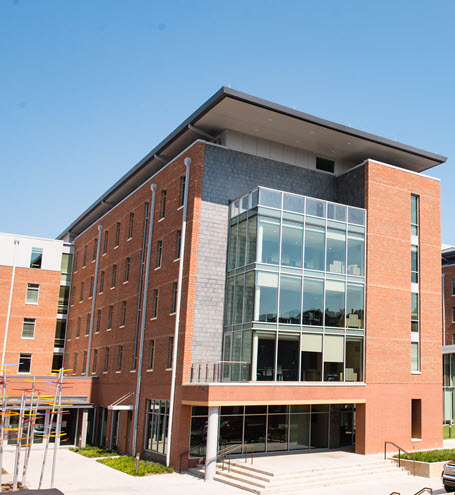 Modern multi-story brick academic buildings with large glass entrances and a street with parked cars in front.