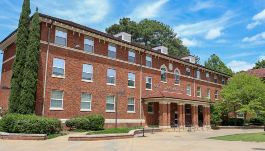 Wide view of a multi-story red brick school building with arched windows and a central entrance, fronted by a large brick-and-concrete courtyard.