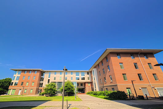 Wide walkway between modern brick-and-glass campus buildings, leading toward a distant domed structure under a cloudy sky.