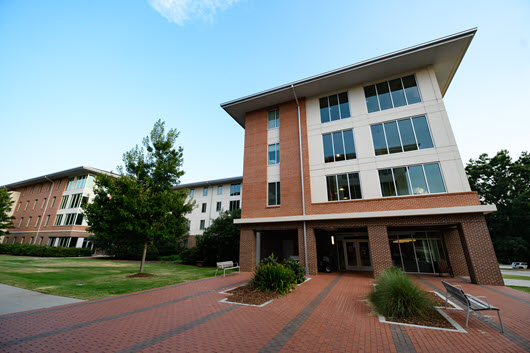 Wide walkway between modern brick-and-glass campus buildings, leading toward a distant domed structure under a cloudy sky.