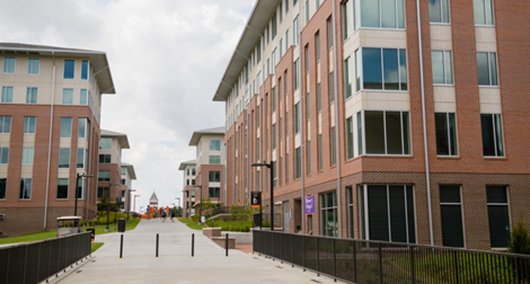 Wide walkway between modern brick-and-glass campus buildings, leading toward a distant domed structure under a cloudy sky.
