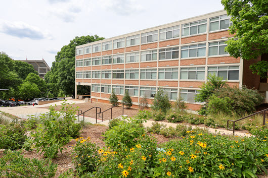 Exterior view of Benet Hall entrance with brick facade, shaded ramp, glass doors, and trees framing the walkway.